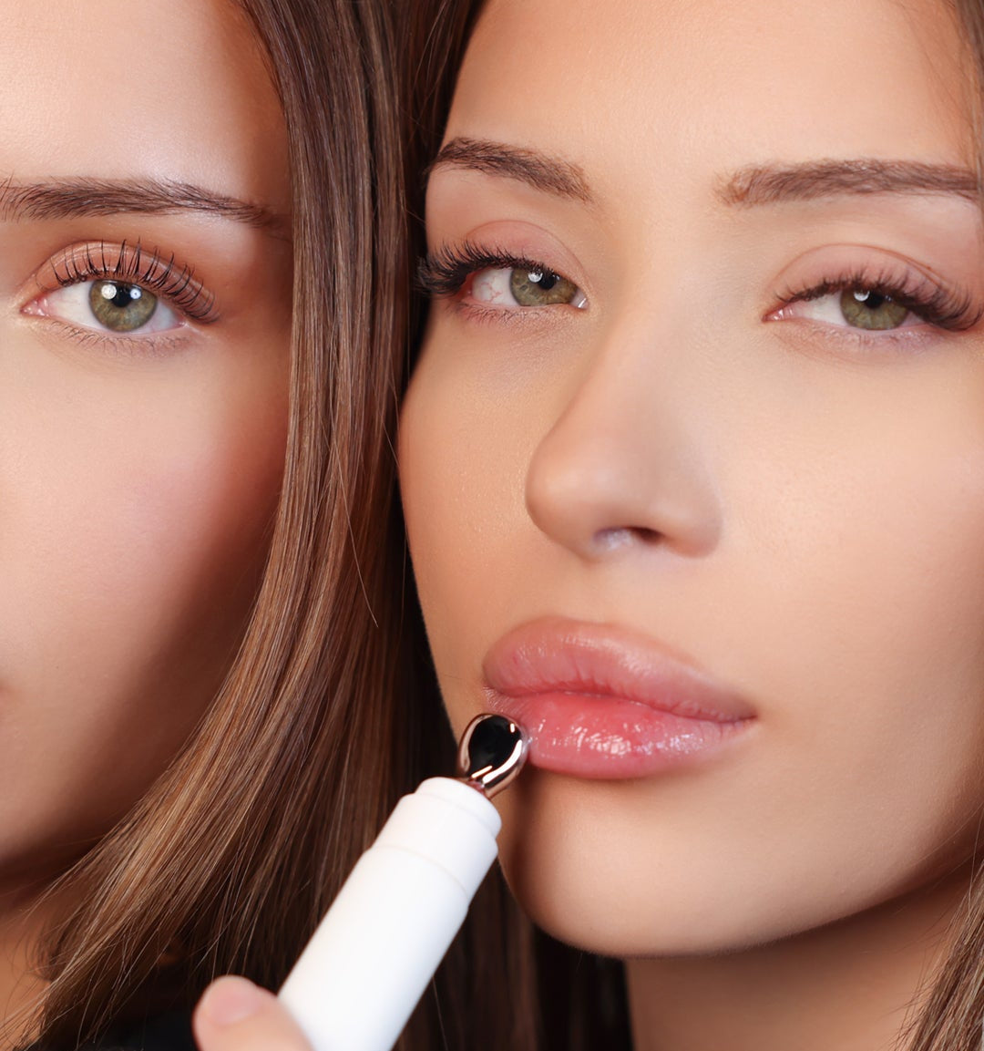 Close-up of a woman holding a white handheld beauty device against her lower lip, with another woman's face partially visible in the background.